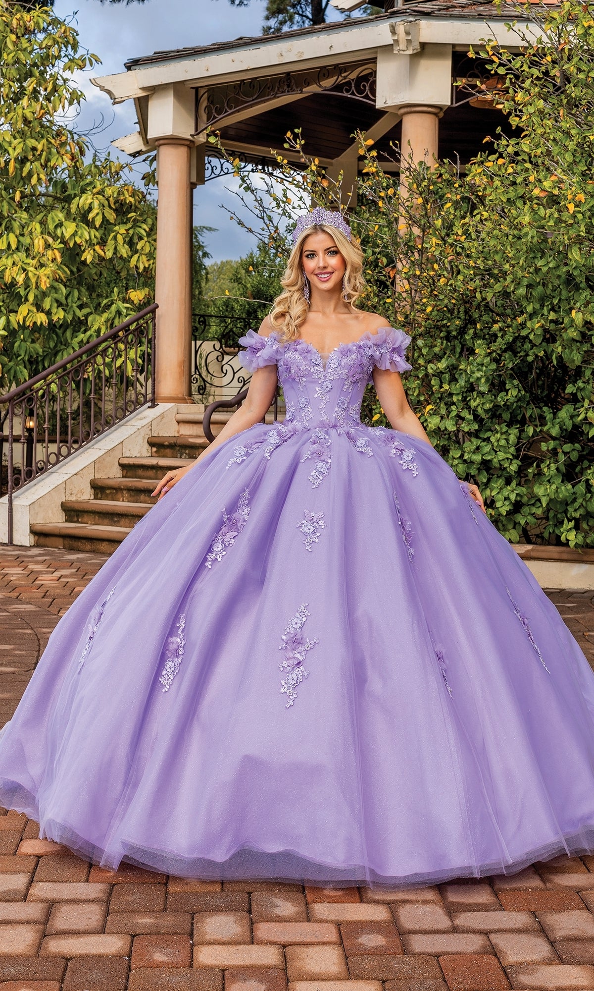 A woman wears the Quinceanera Dress 1870 By Dancing Queen, featuring a lilac hue, floral embellishments, sweetheart neckline, and matching headpiece, as she stands outdoors before a staircase and greenery.